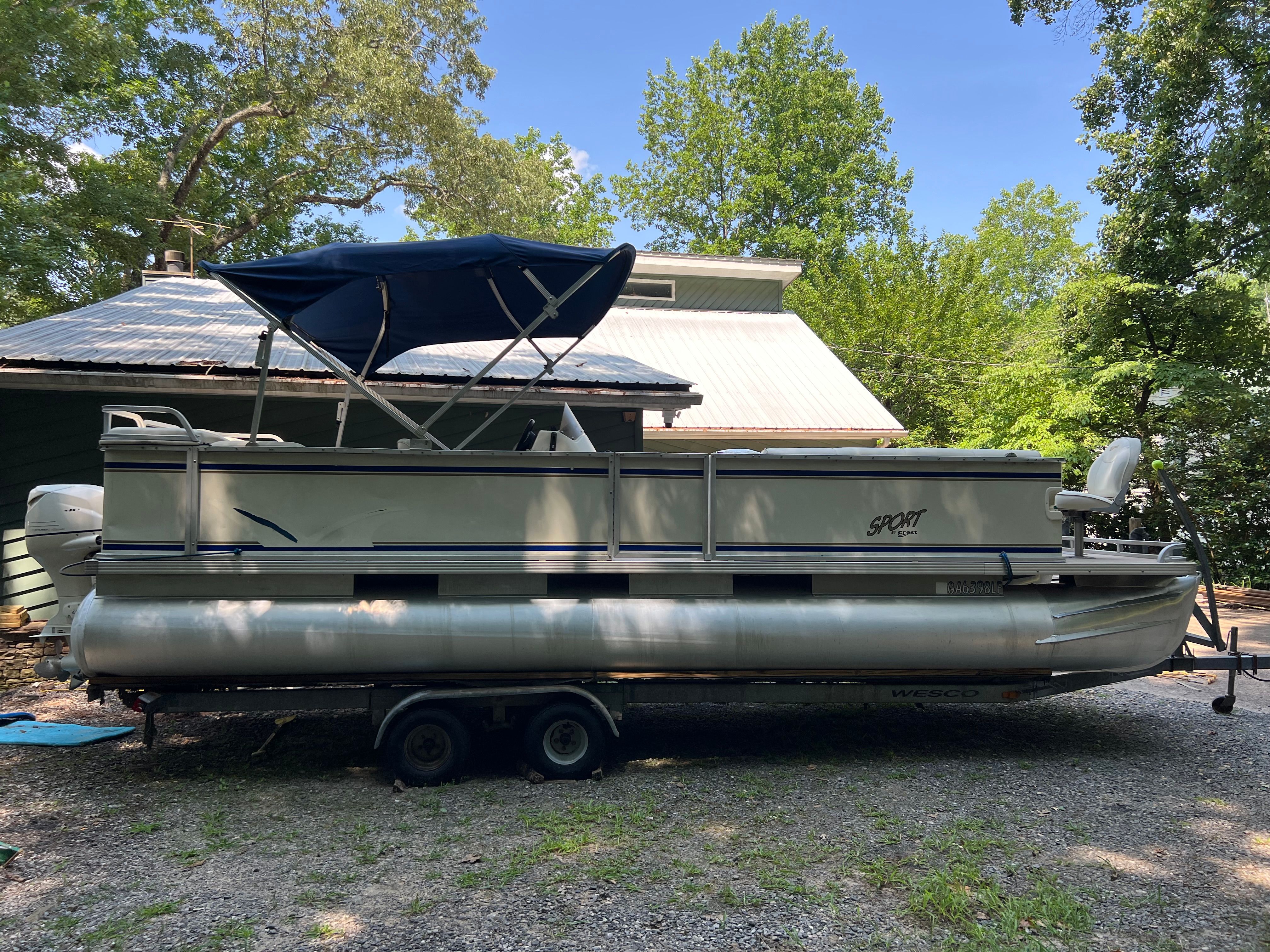 A 2003 Crest 24’ Sport pontoon boat on a trailer, surrounded by trees and a building.