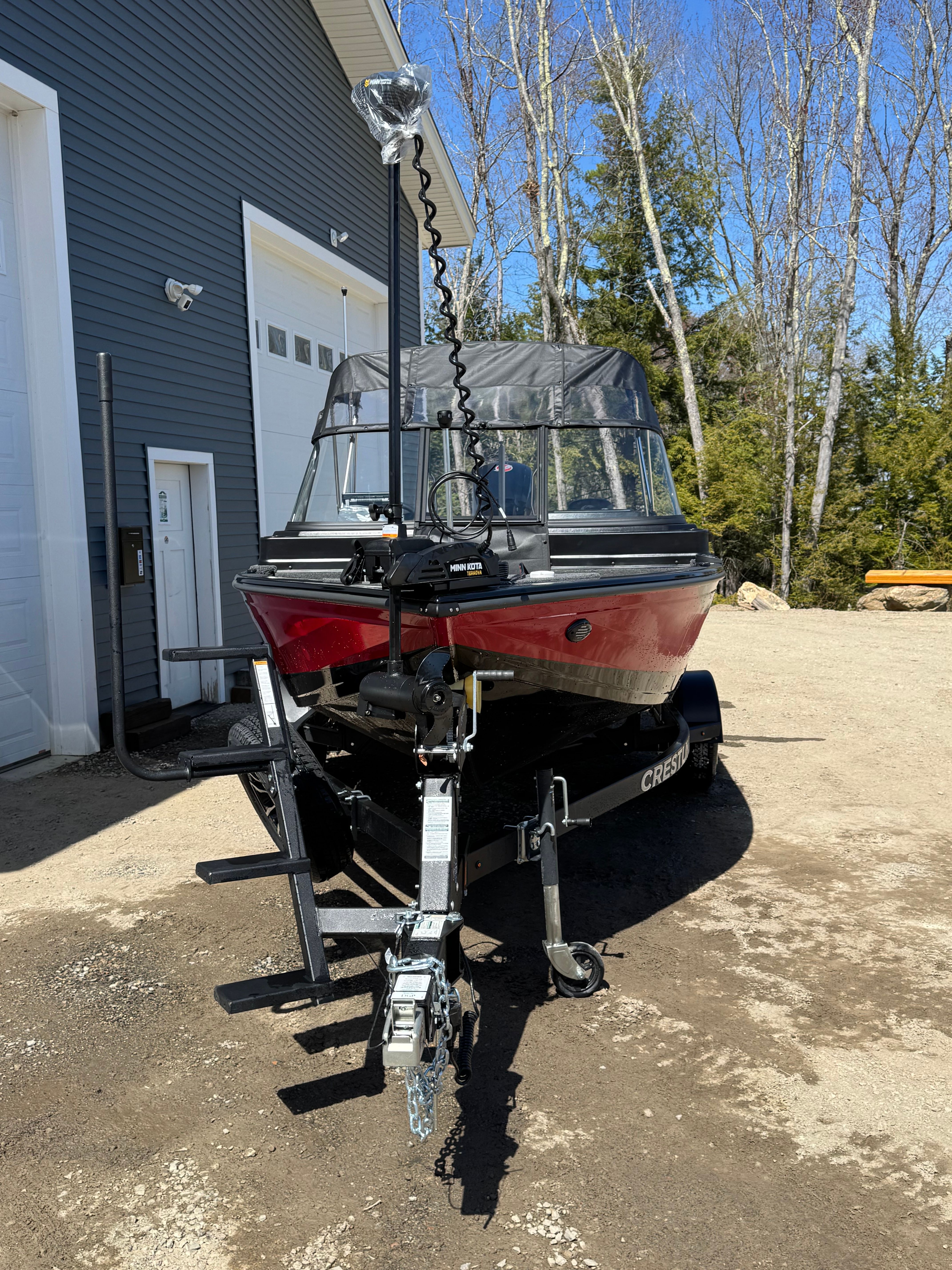 A red boat on a trailer in front of a garage, surrounded by trees.