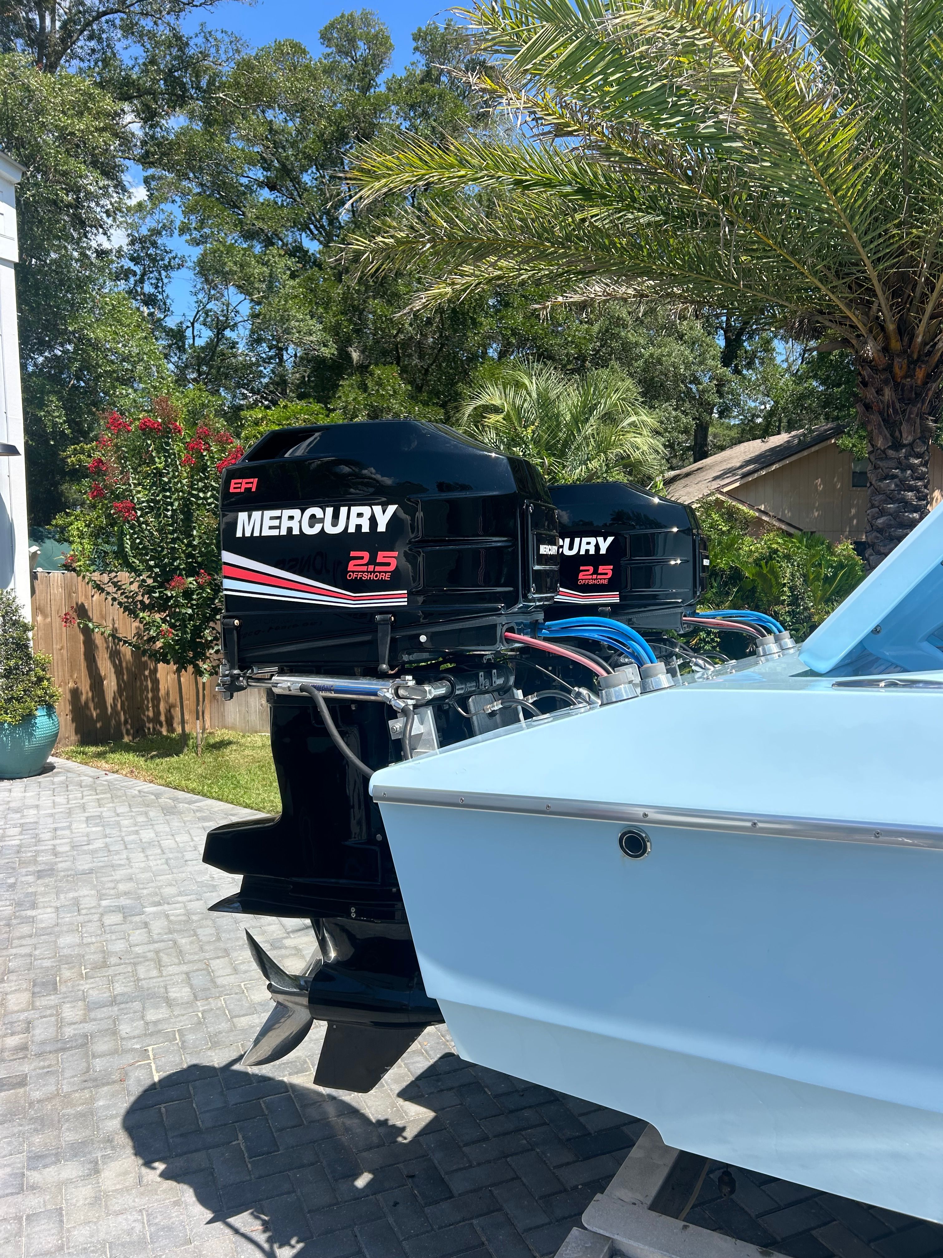 Two Mercury 2.5 outboard engines on a blue boat, surrounded by palm trees and a paved yard.