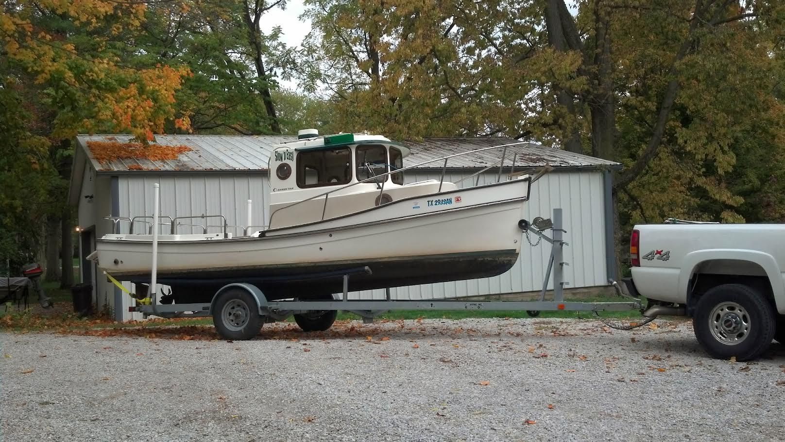 A 2007 Ranger Tugs R-21 EC on a trailer, parked in front of a shed with autumn trees.