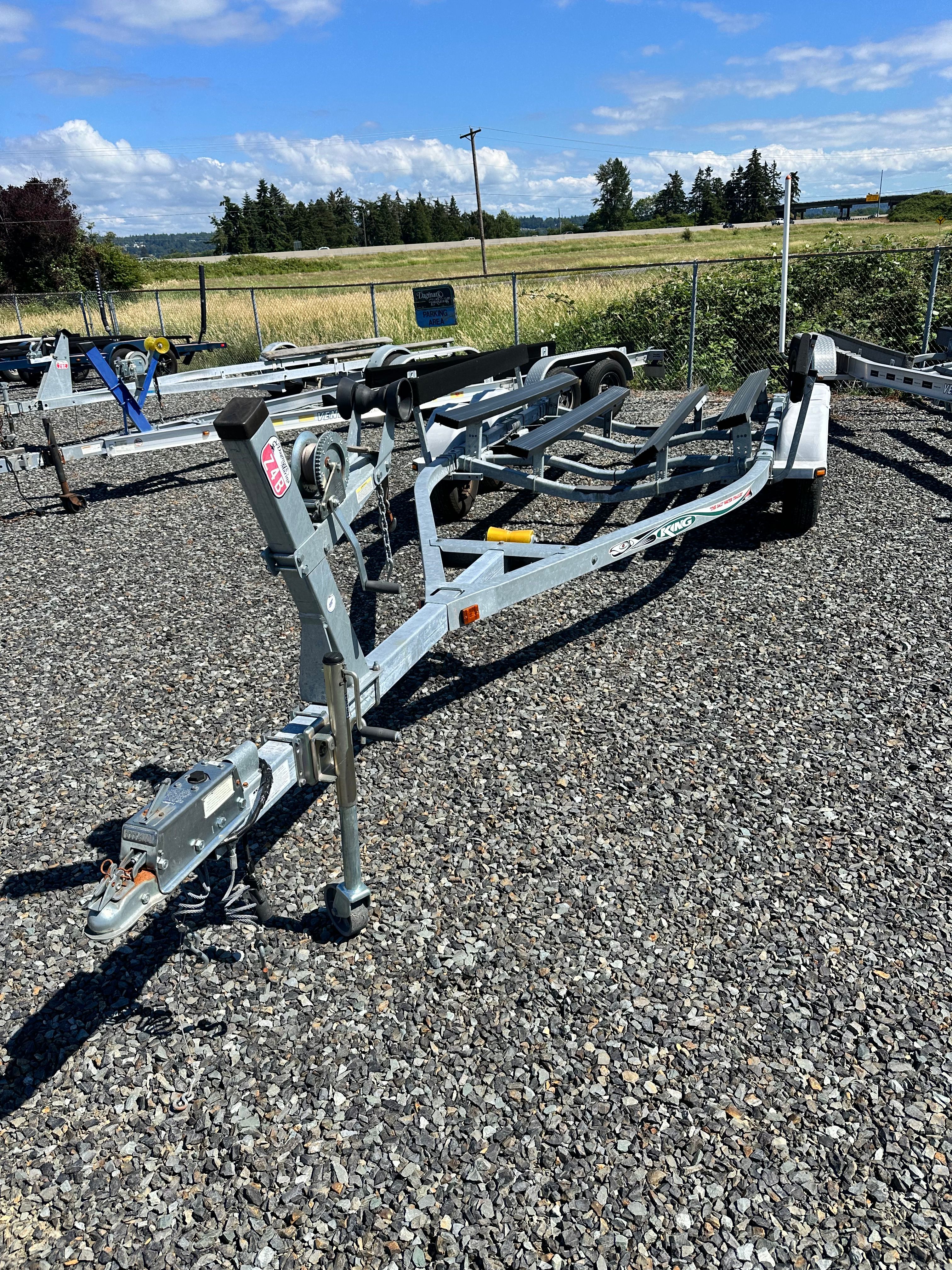 A boat trailer sits on a gravel lot, surrounded by other trailers and a fence.