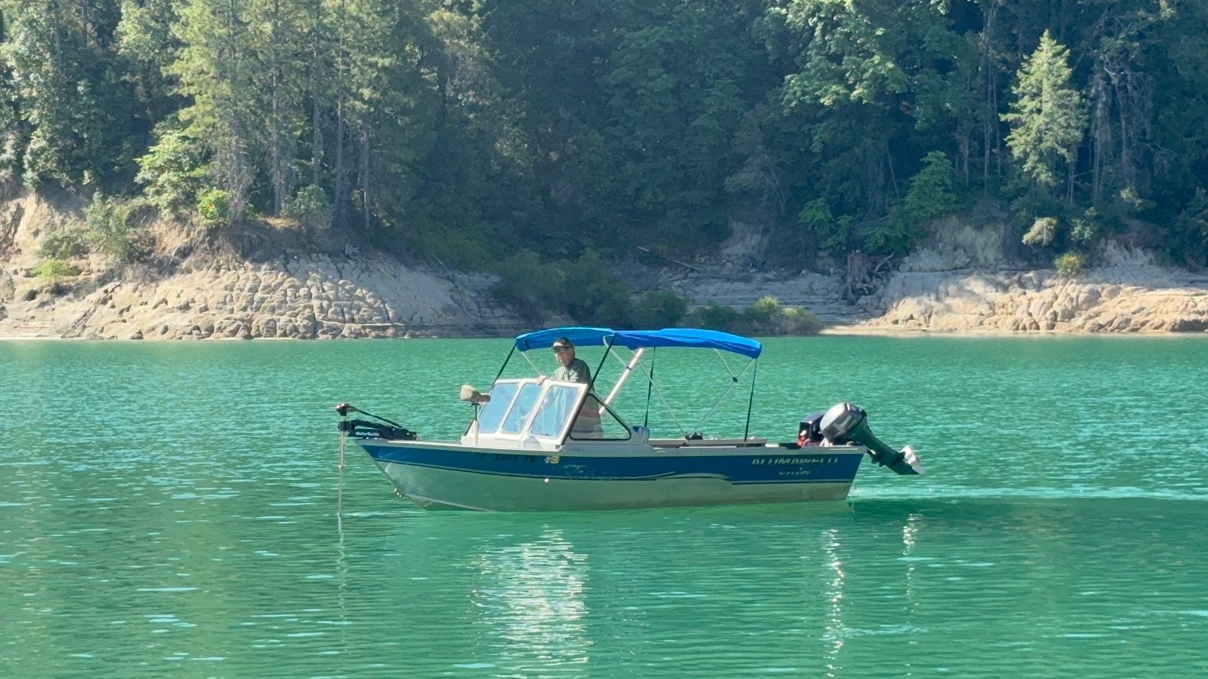 A small motorboat with a blue canopy on a calm lake, surrounded by trees.
