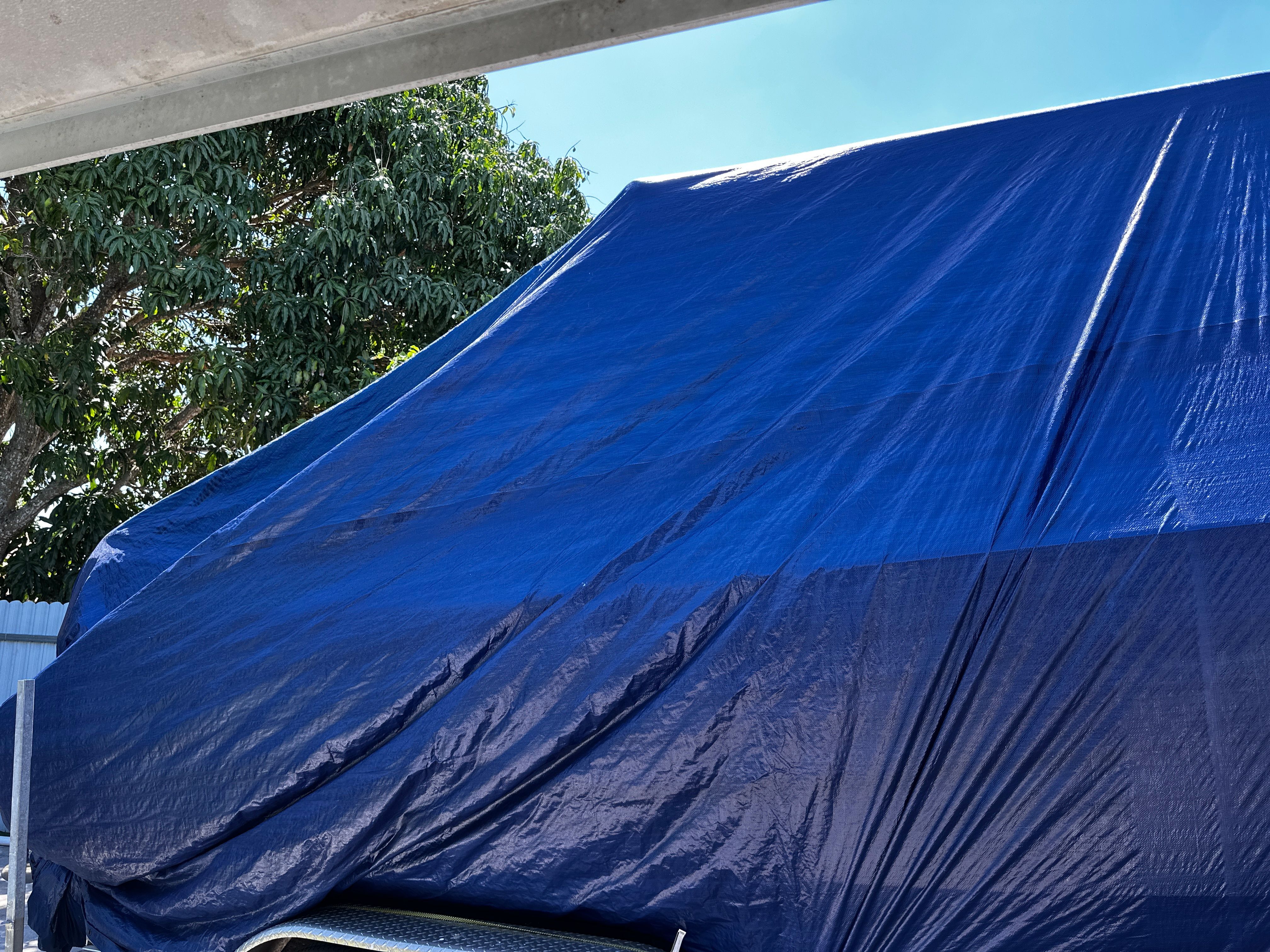 A boat covered with a blue tarp is parked under a tree.