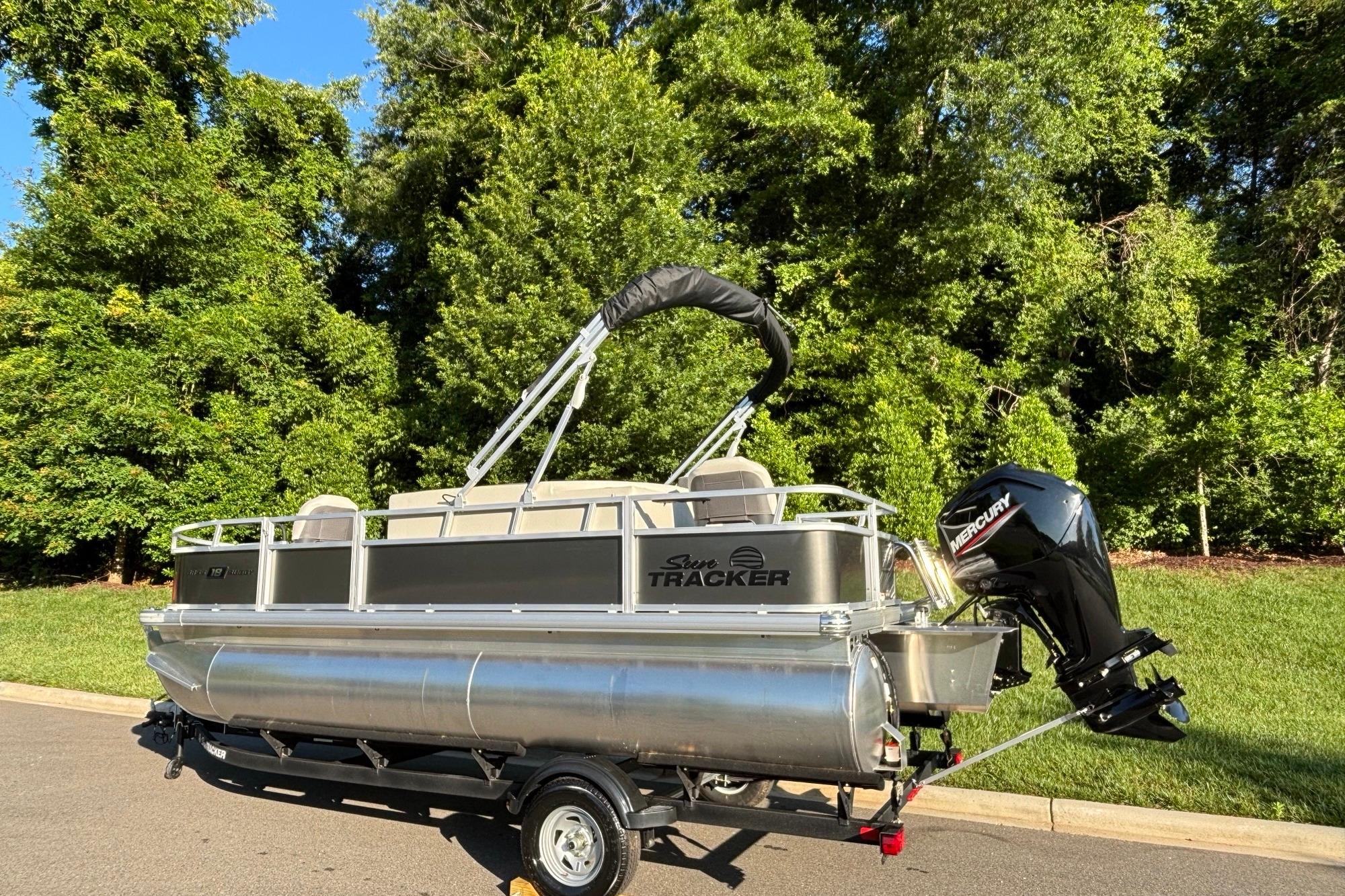 2026 Sun Tracker Bass Buggy 18 DLX pontoon boat on trailer, parked near lush greenery.