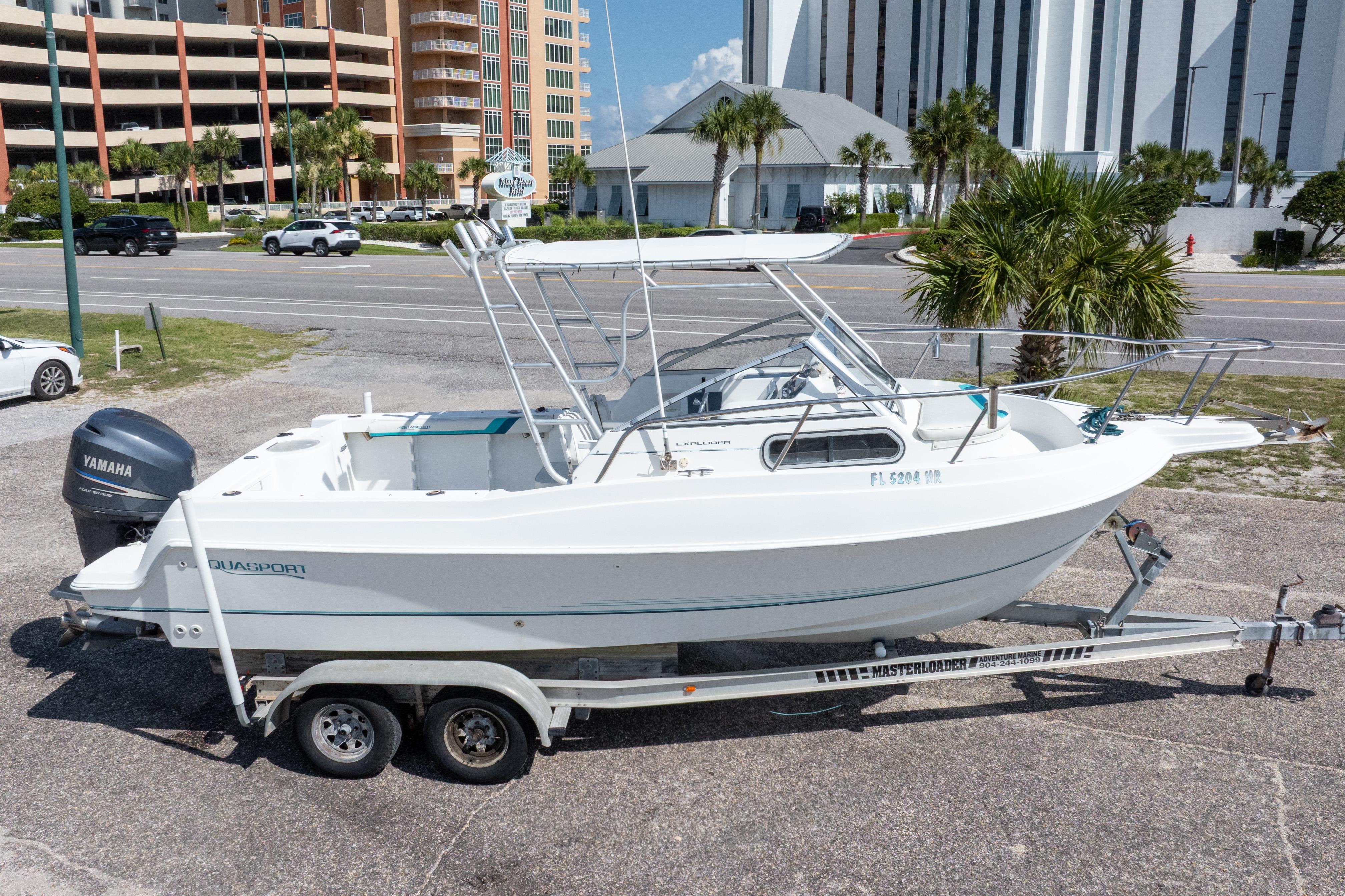 1999 Aquasport 225 Explorer boat on trailer, parked near road with buildings in background.