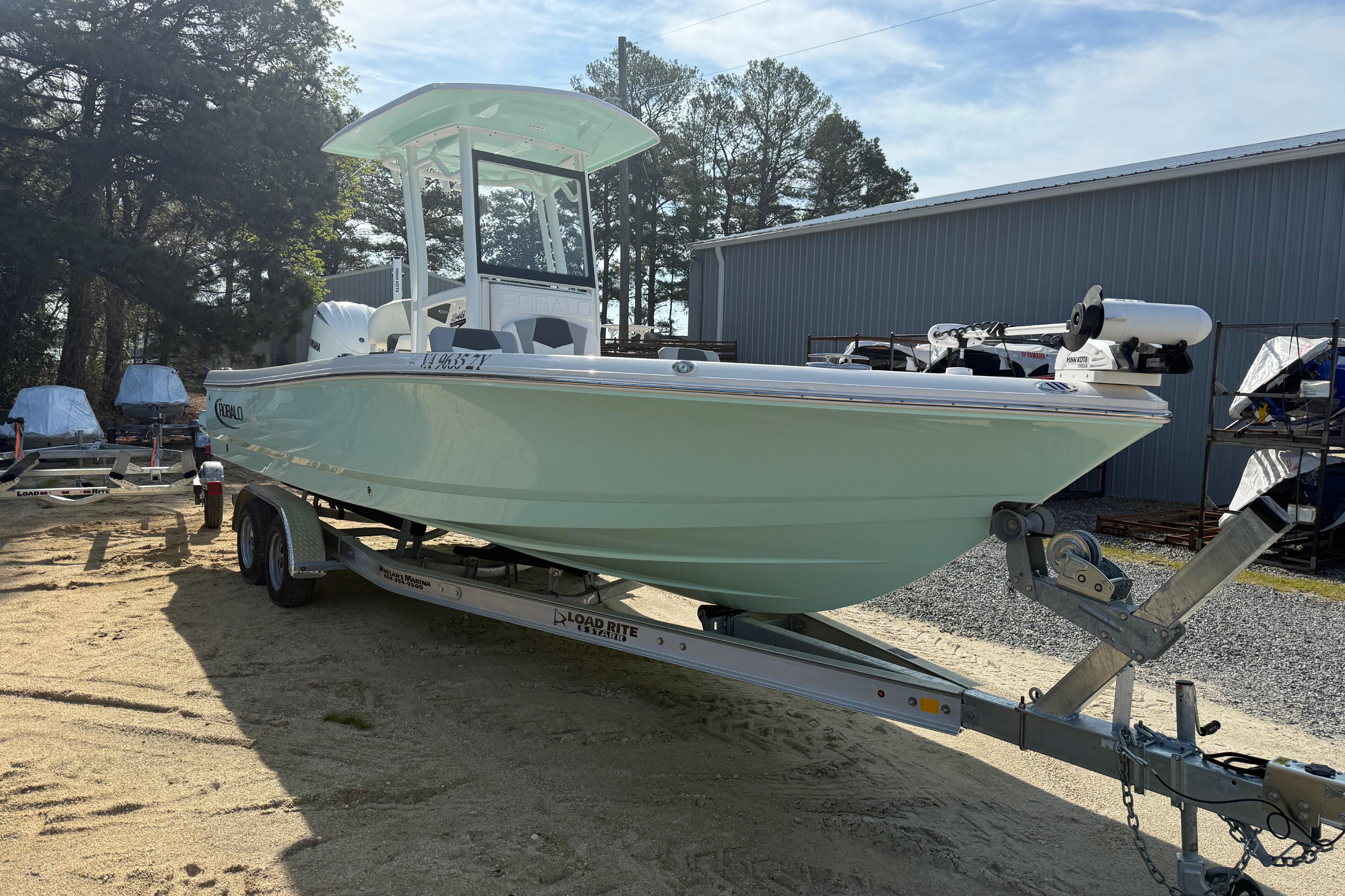 2023 Robalo 246 Cayman boat on trailer, parked outdoors near trees and a building.