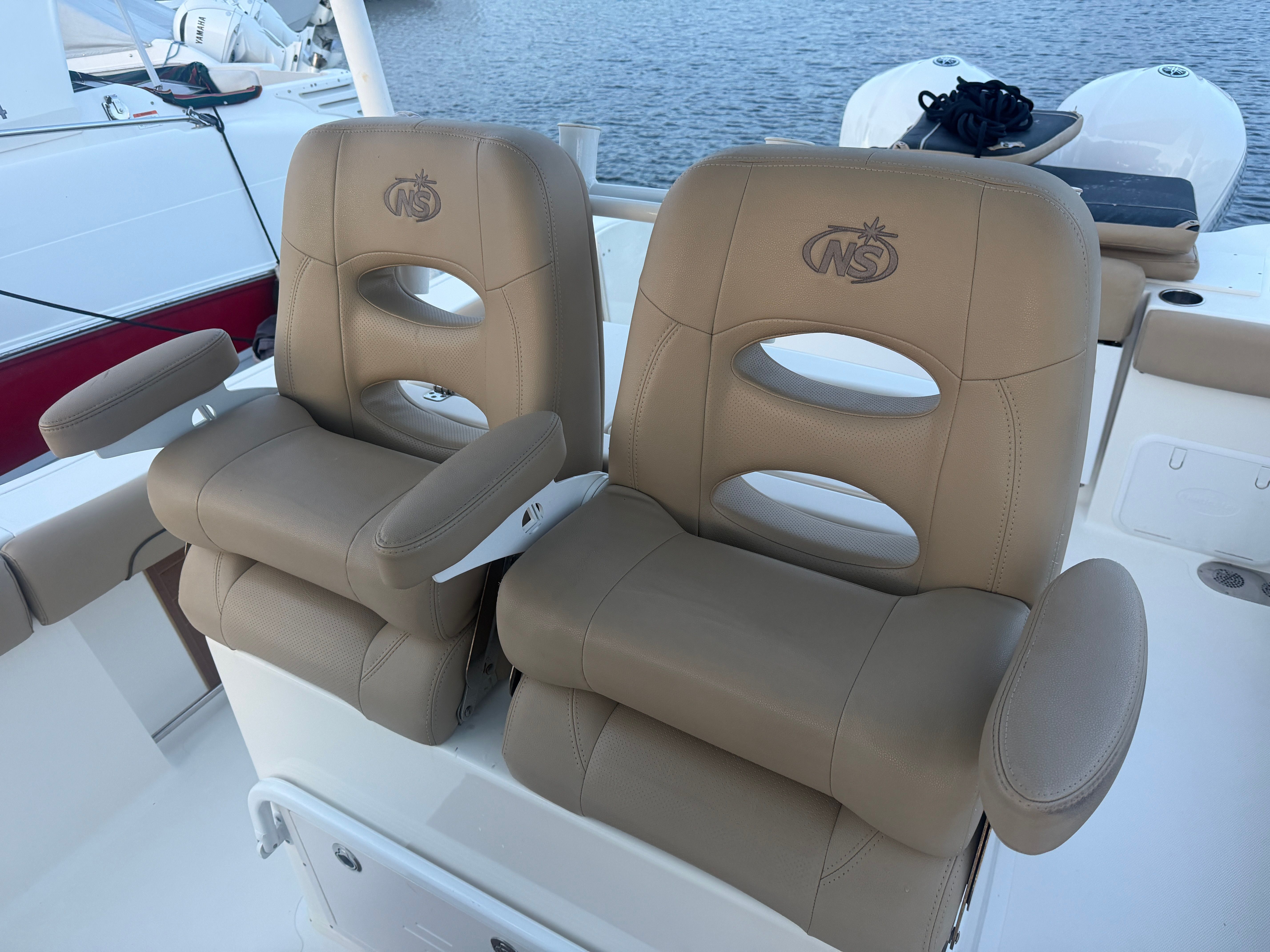 Two beige captain's chairs on a boat with armrests and NS logo, set against calm waters.