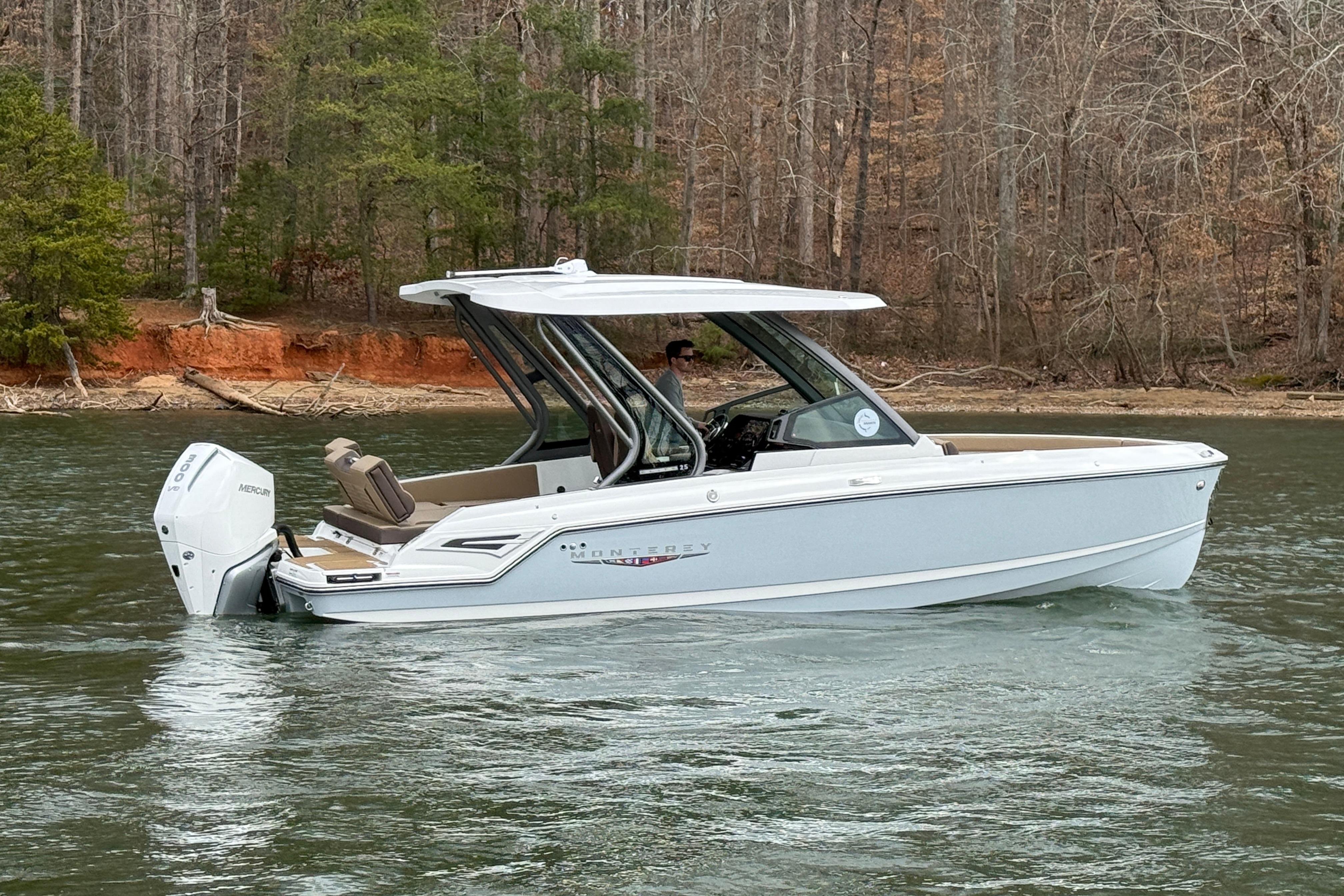 2026 Monterey Elite 25 OB boat cruising on a serene lake with wooded shoreline.