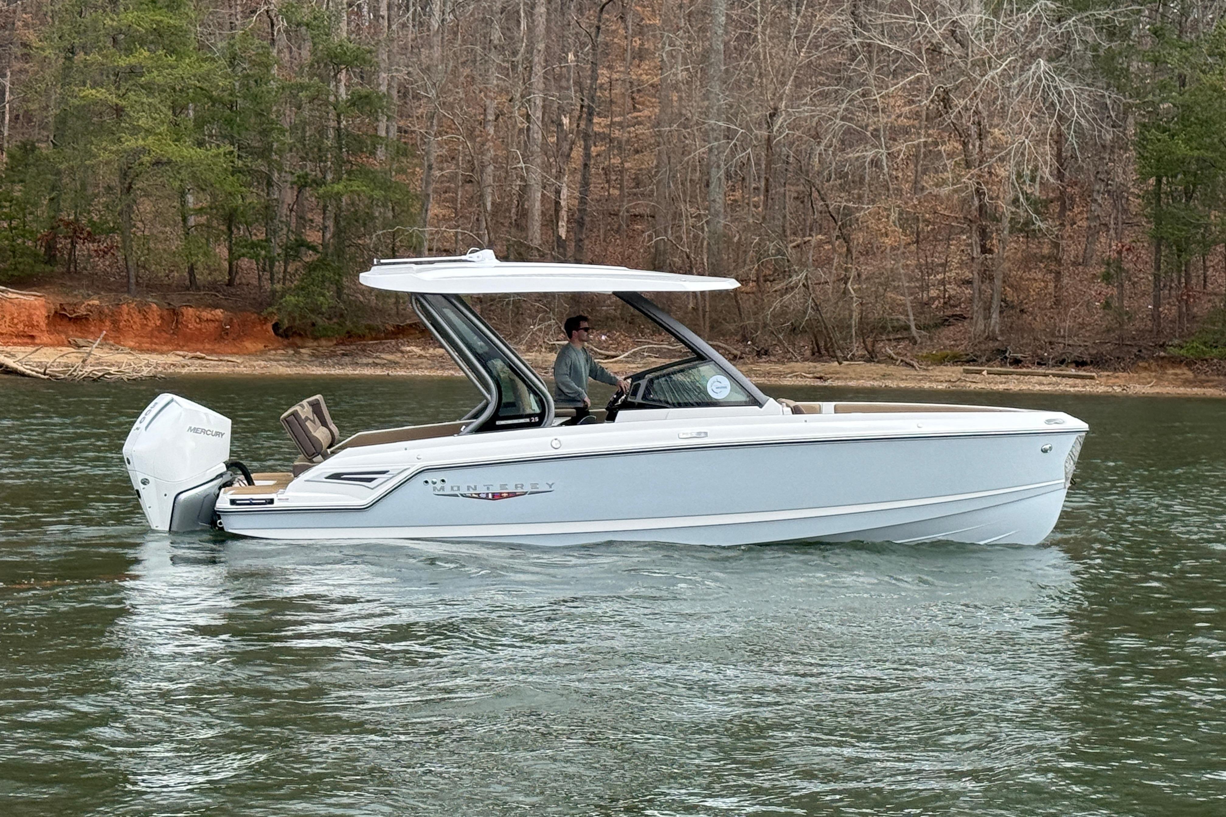 2026 Monterey Elite 25 OB boat cruising on a serene lake with wooded shoreline.