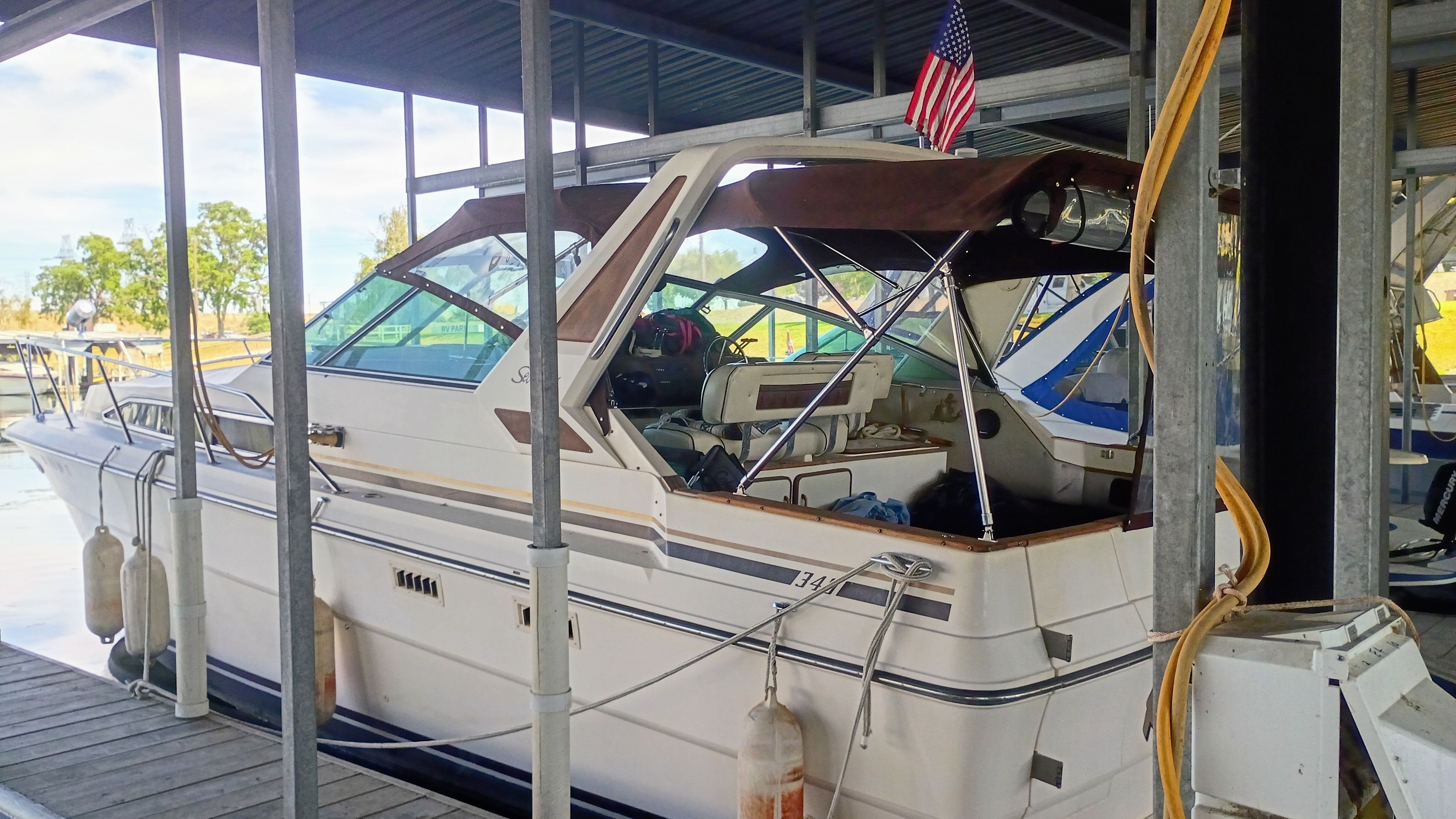 A 1986 Sea Ray Sundancer docked under a covered area. It features a brown canopy and an American flag.