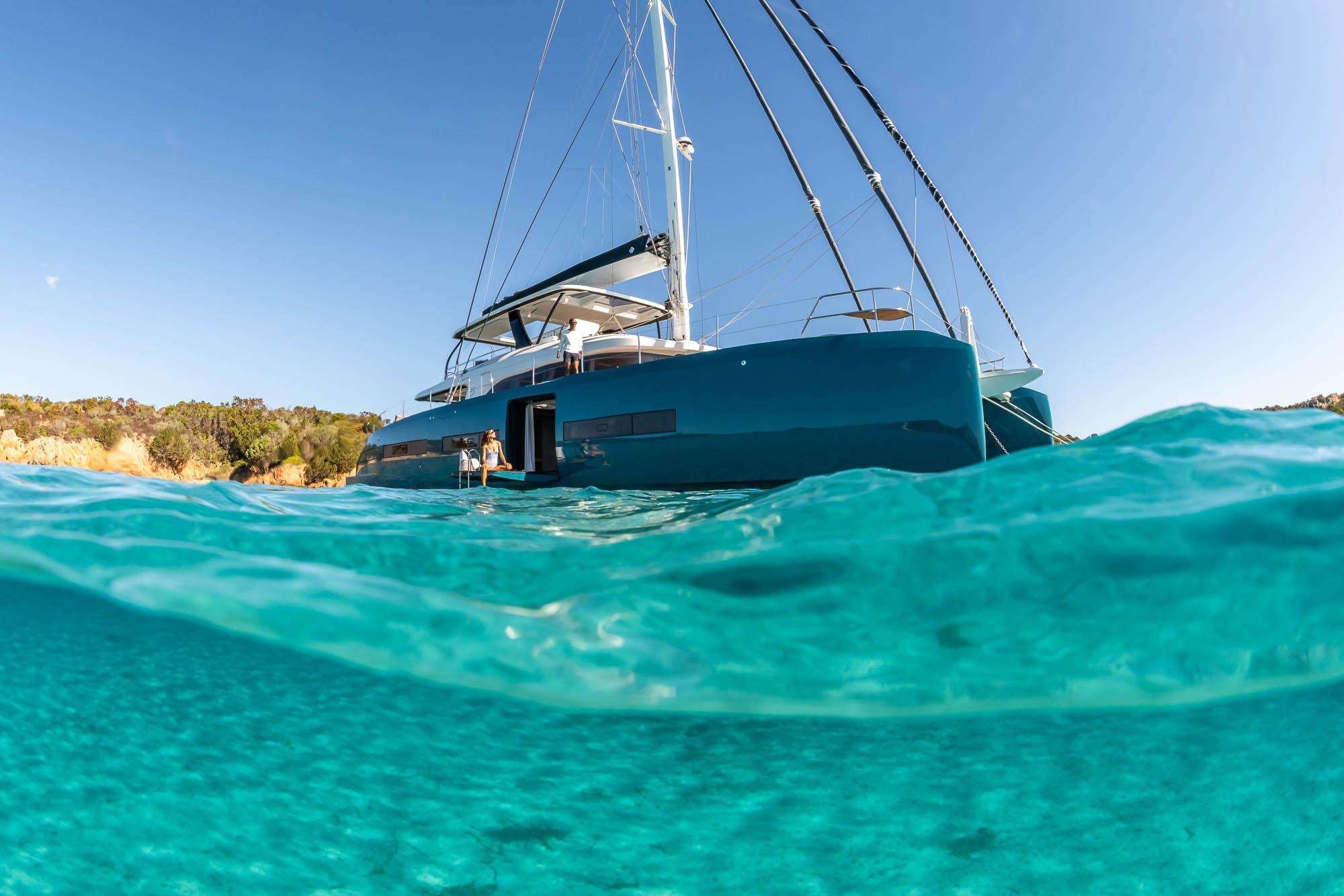 2026 Lagoon Eighty 2 yacht in clear turquoise water, sunny day, coastal backdrop.
