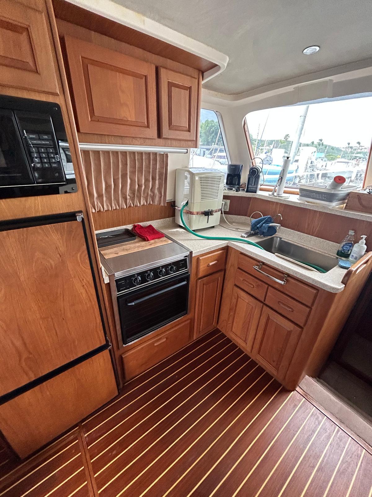 1999 Mainship 350 Trawler kitchen with wooden cabinetry, appliances, and a window view.