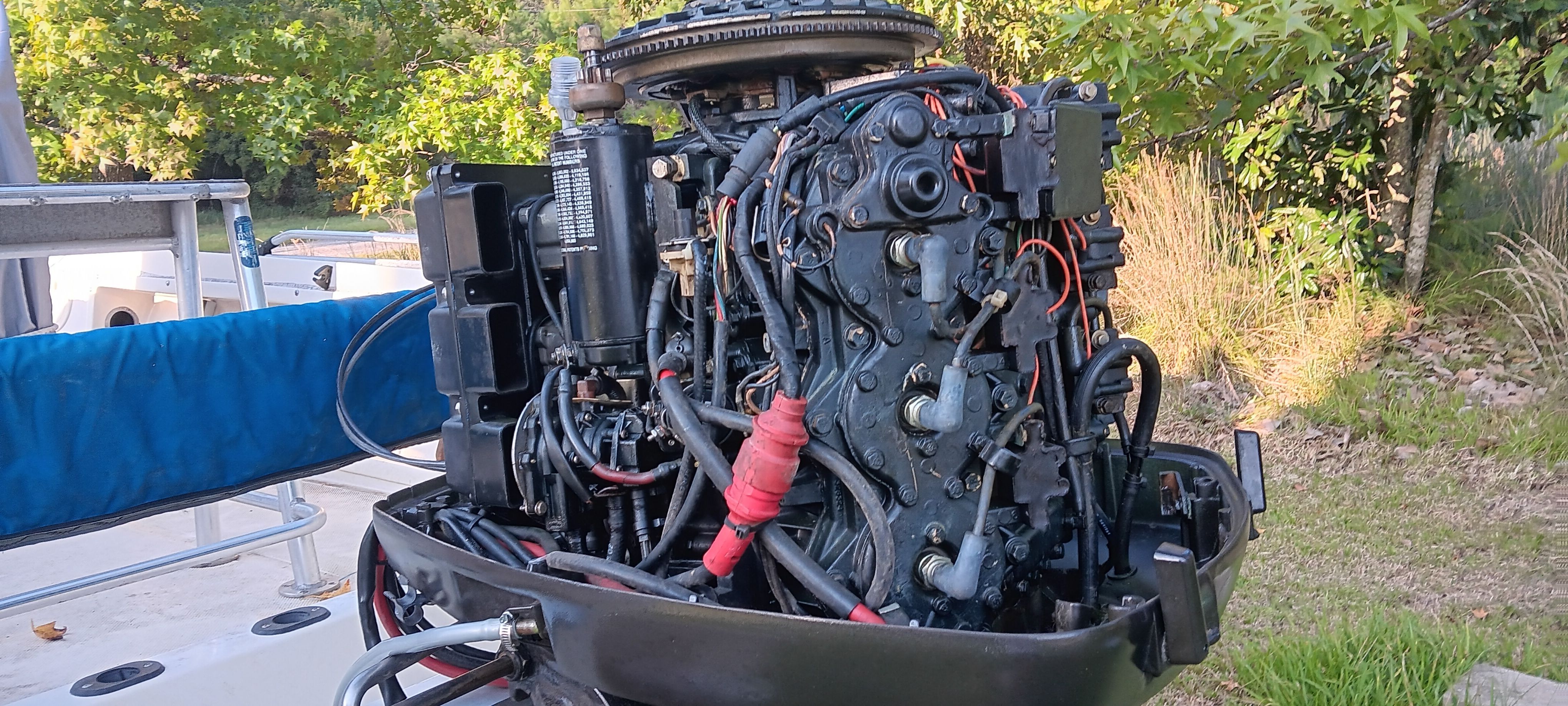 An exposed boat engine with visible wires and components, surrounded by trees and grass.