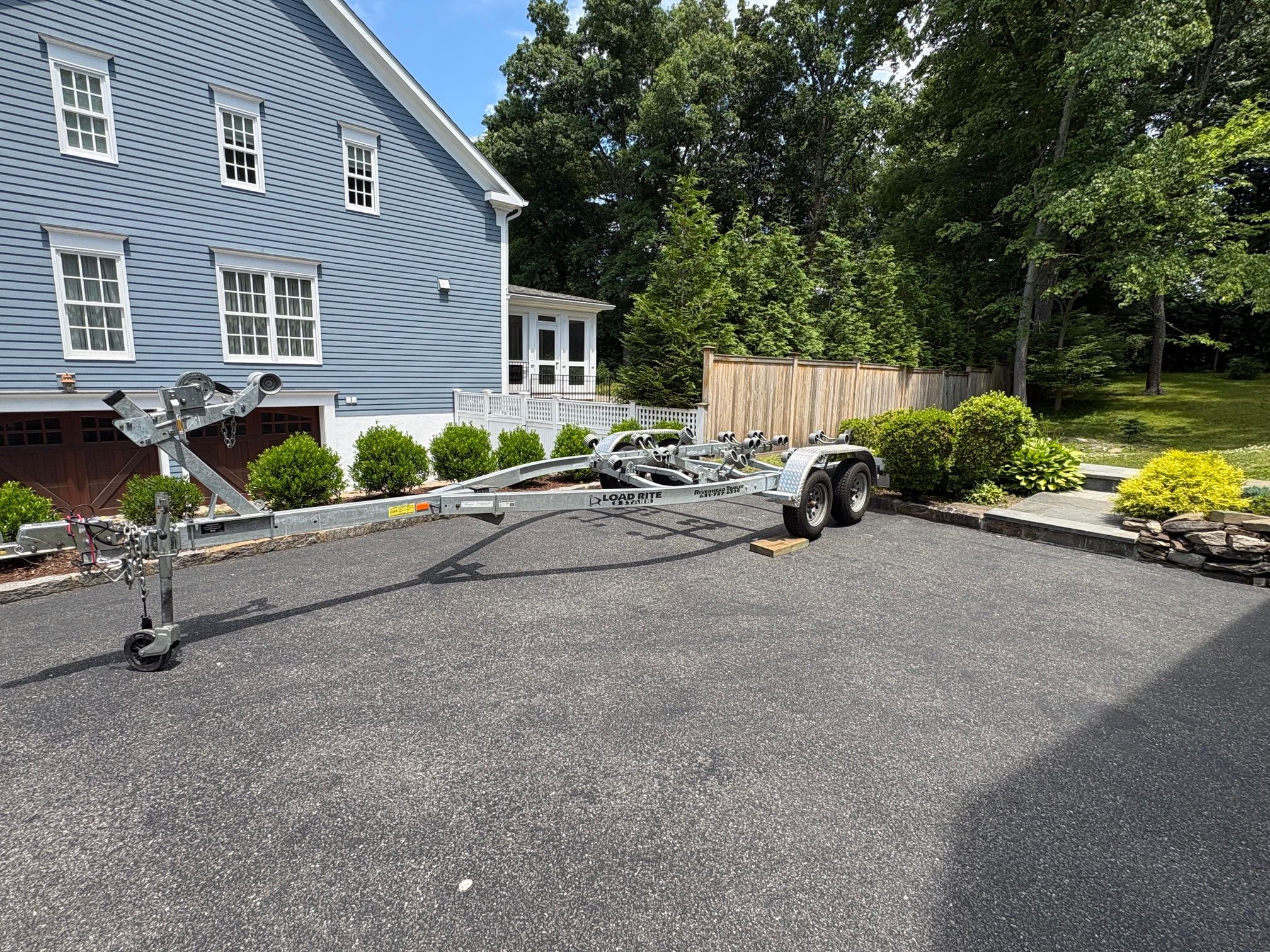 A boat trailer is parked on a paved driveway in front of a blue house with white window frames.