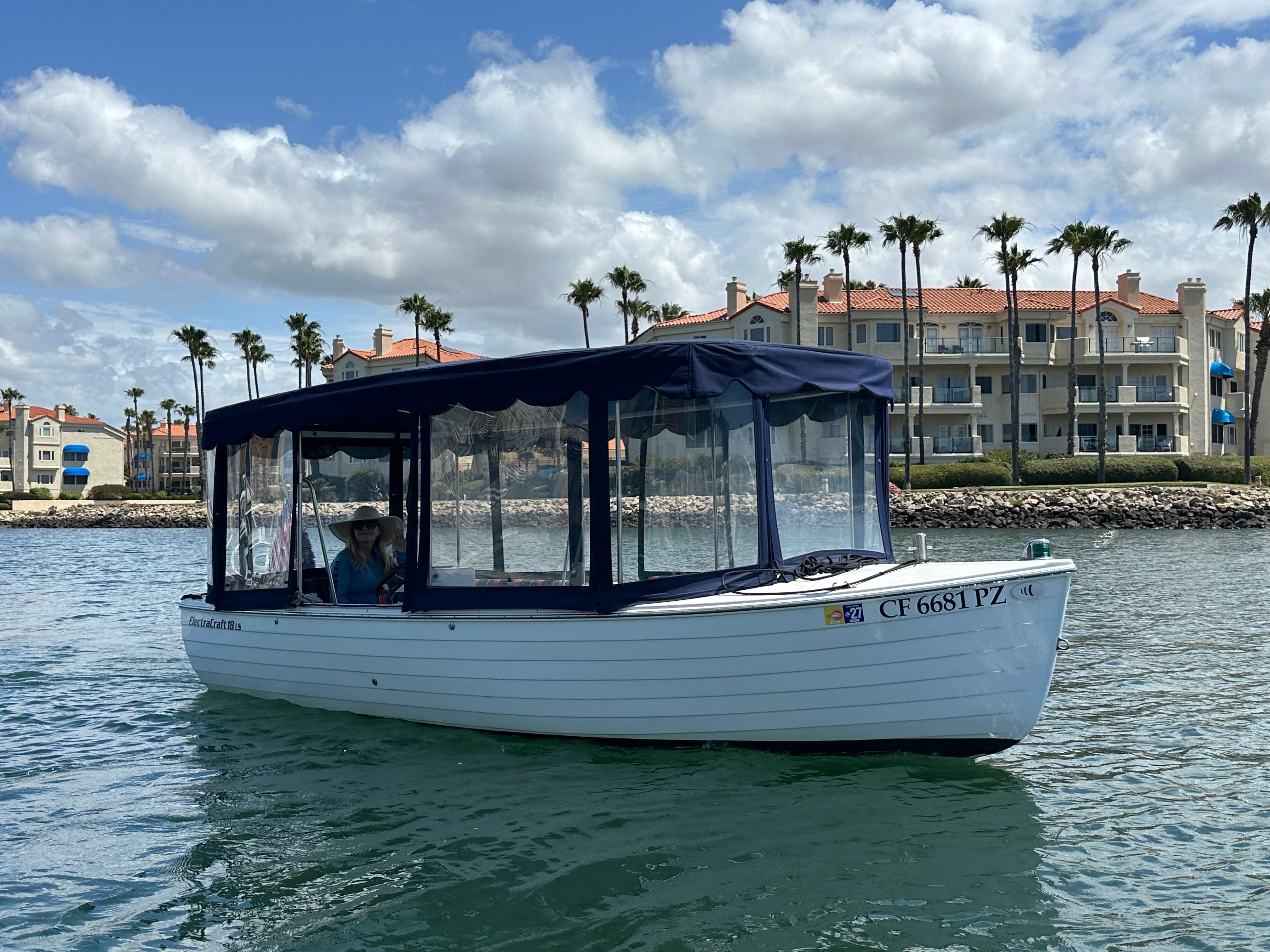 A white boat with a blue canopy cruises on calm water near a shoreline with palm trees.