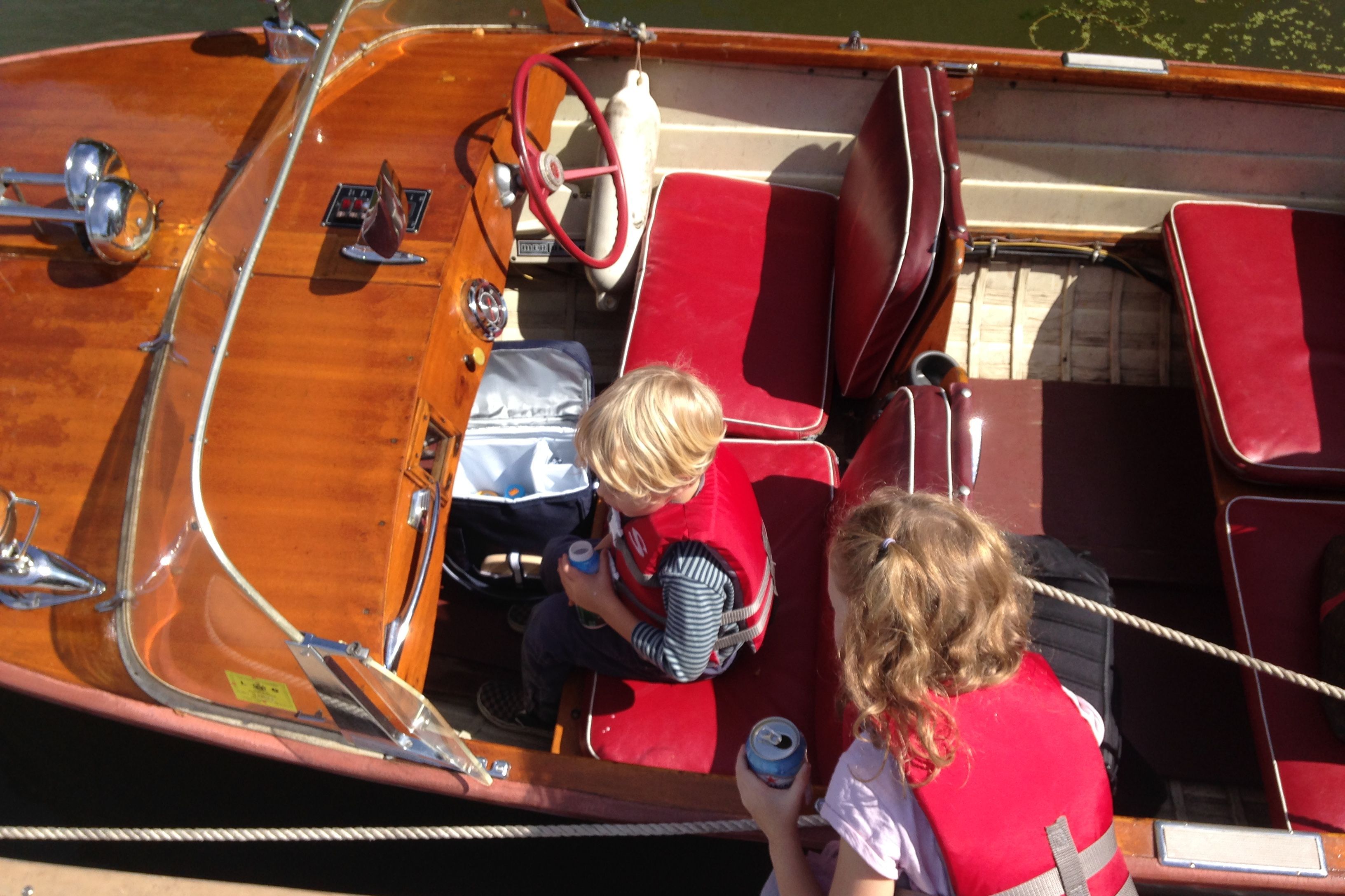 Two children in red life jackets sit in a classic wooden boat with a red steering wheel.