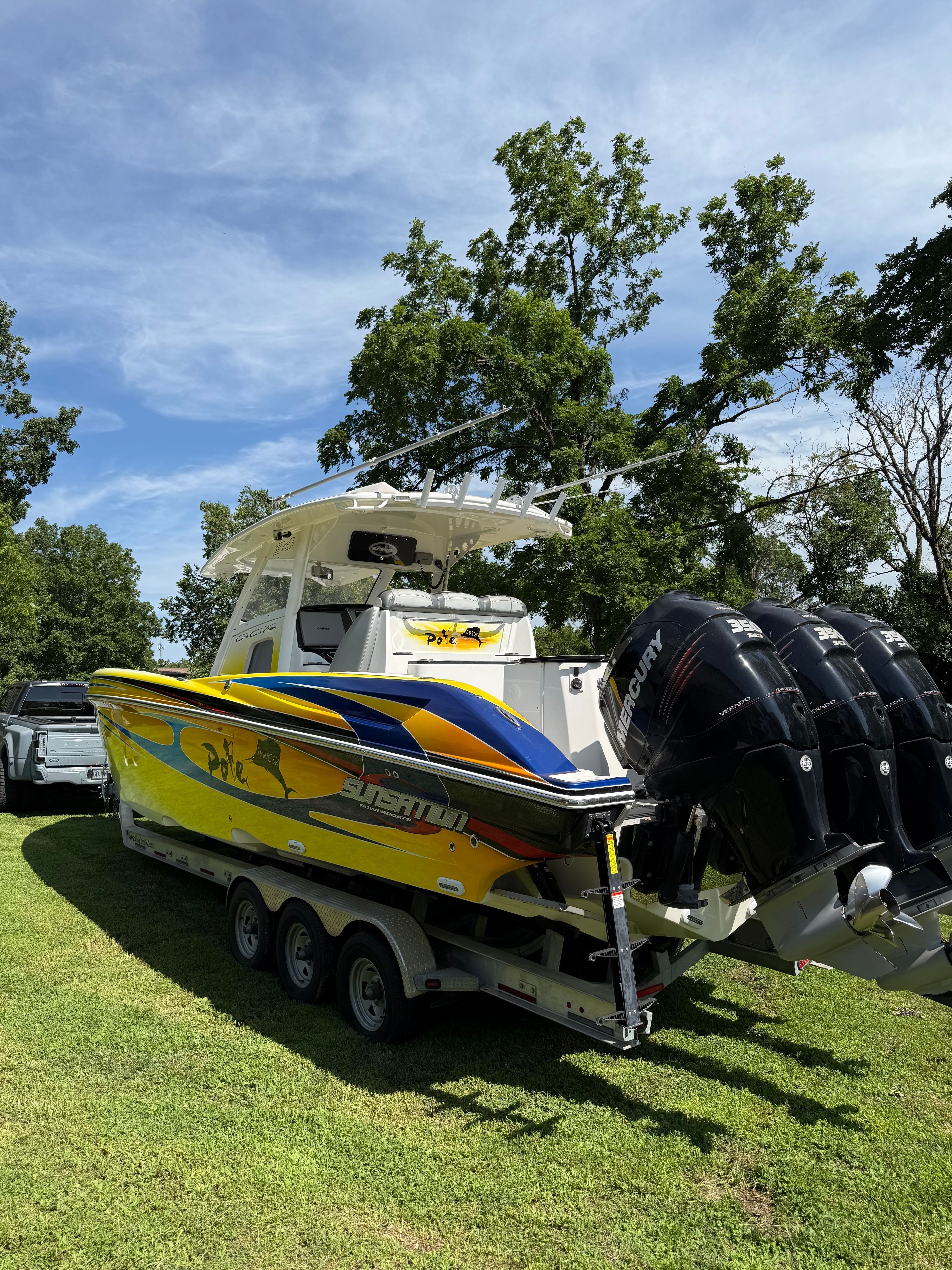 A yellow and blue boat on a trailer with three engines, surrounded by trees.