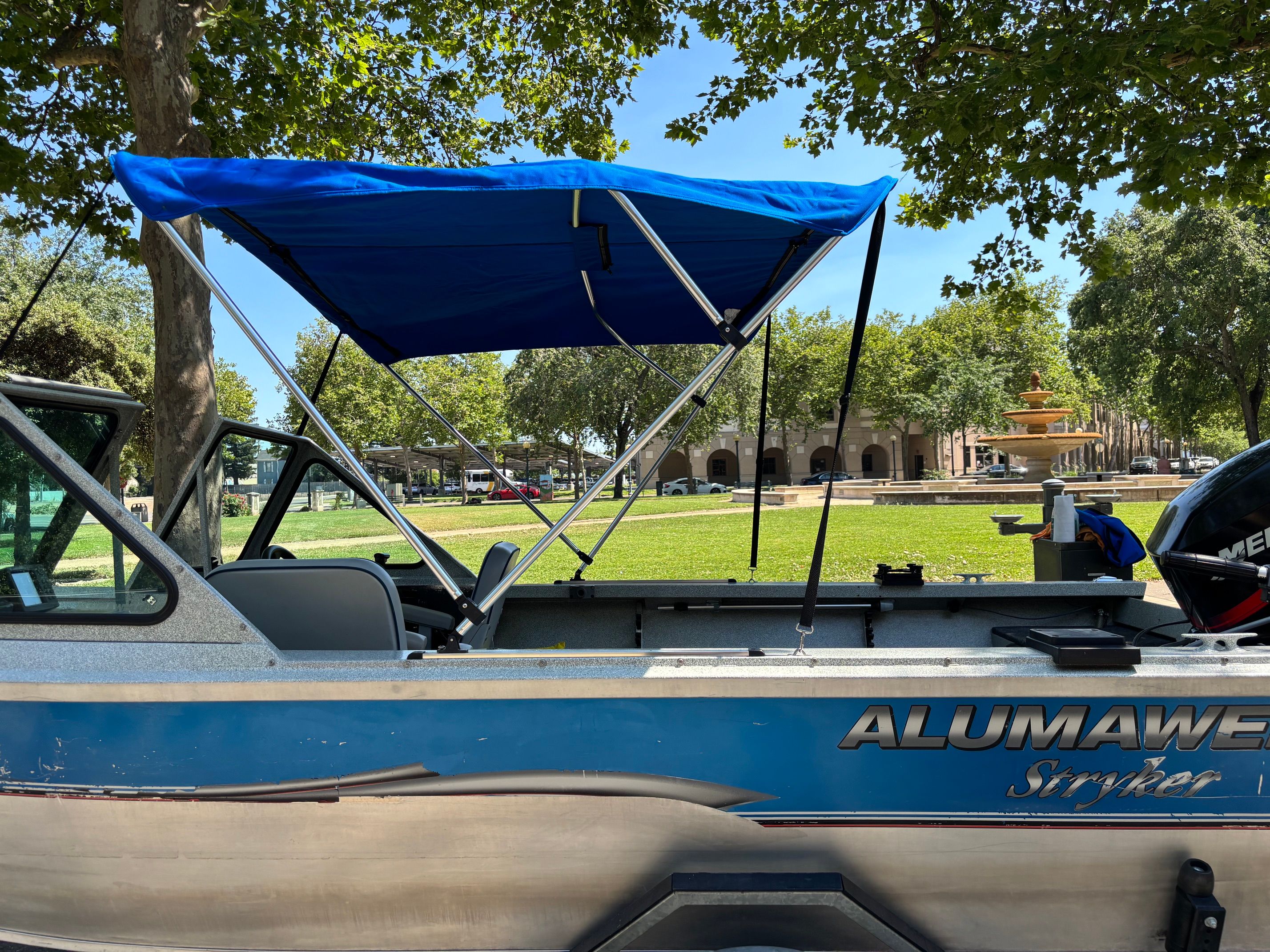 A blue Alumaweld boat with a blue sunshade is on a trailer in a park.