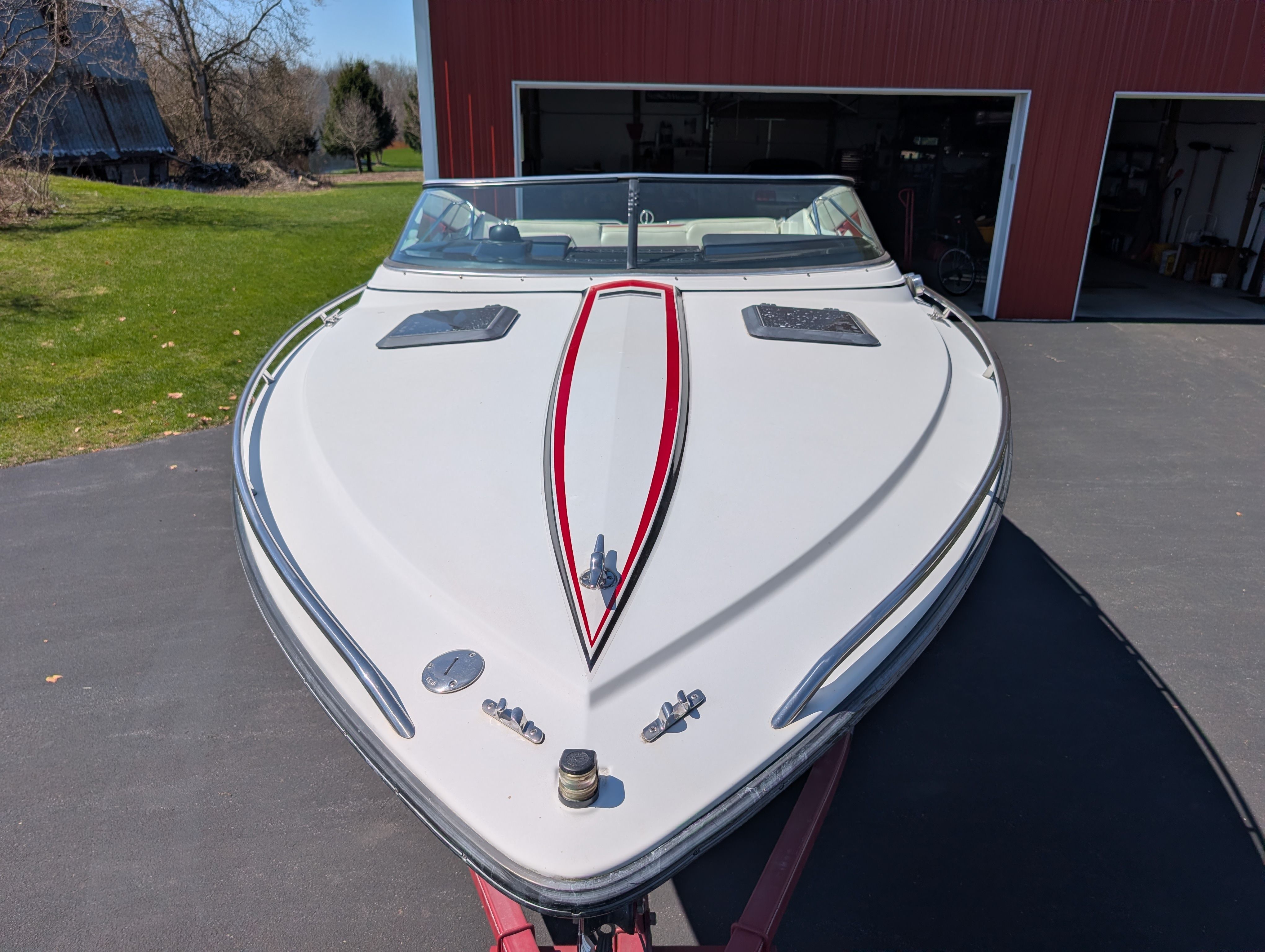 A white boat with a red stripe is on a trailer in front of a barn.