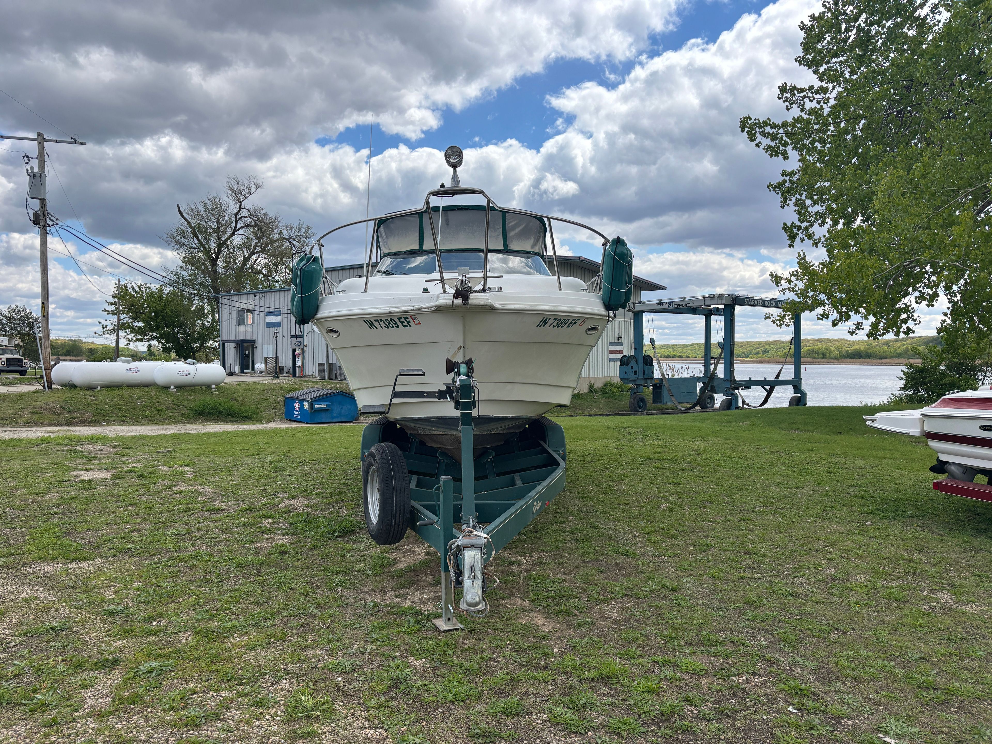A boat on a trailer sits on grass, surrounded by trees and buildings.