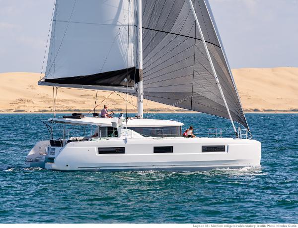 2025 Lagoon 46 ICONIC catamaran sailing on blue ocean near sandy dunes.