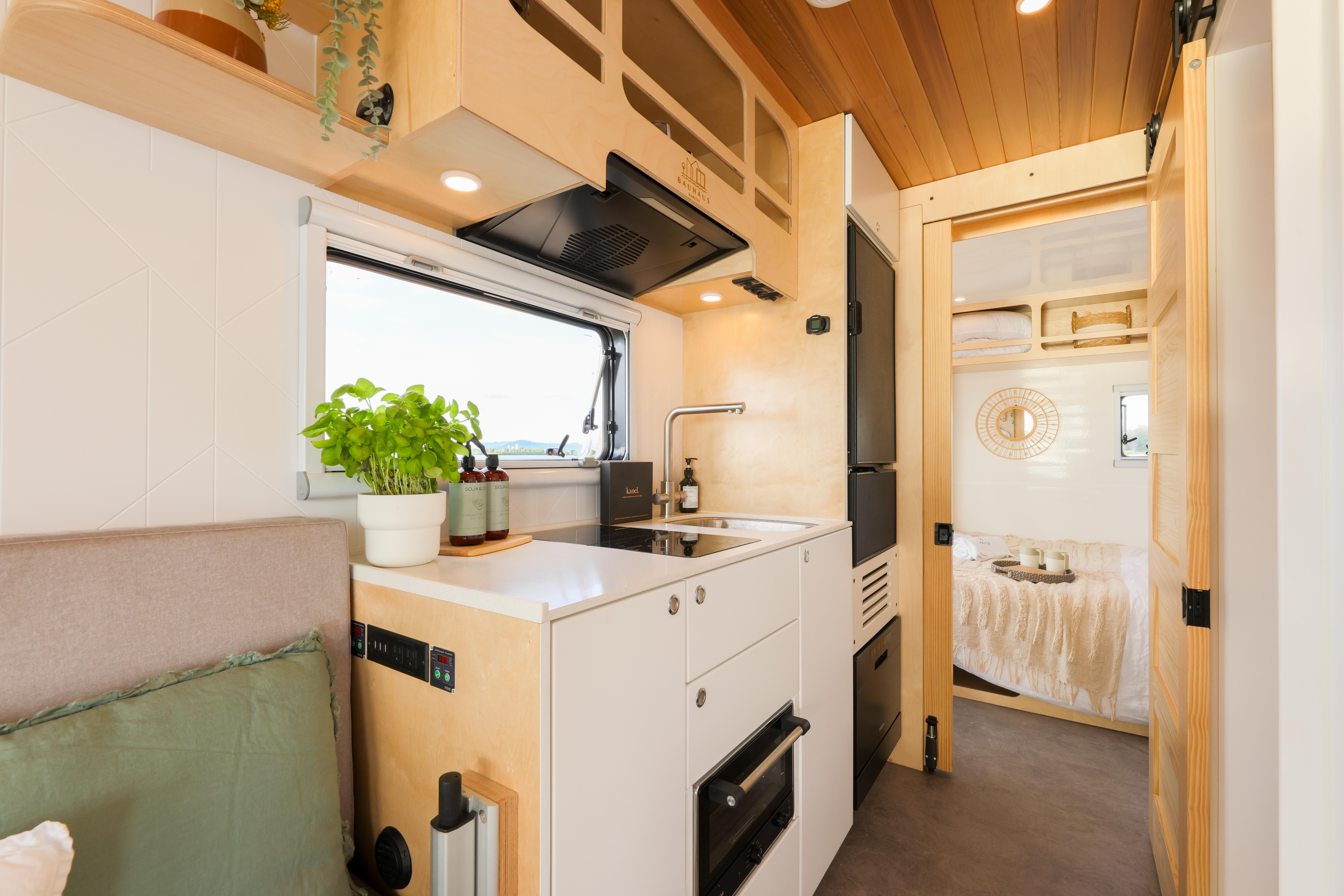Modern camper van kitchen interior with sleek cabinetry, compact appliances, and cozy bedroom view.