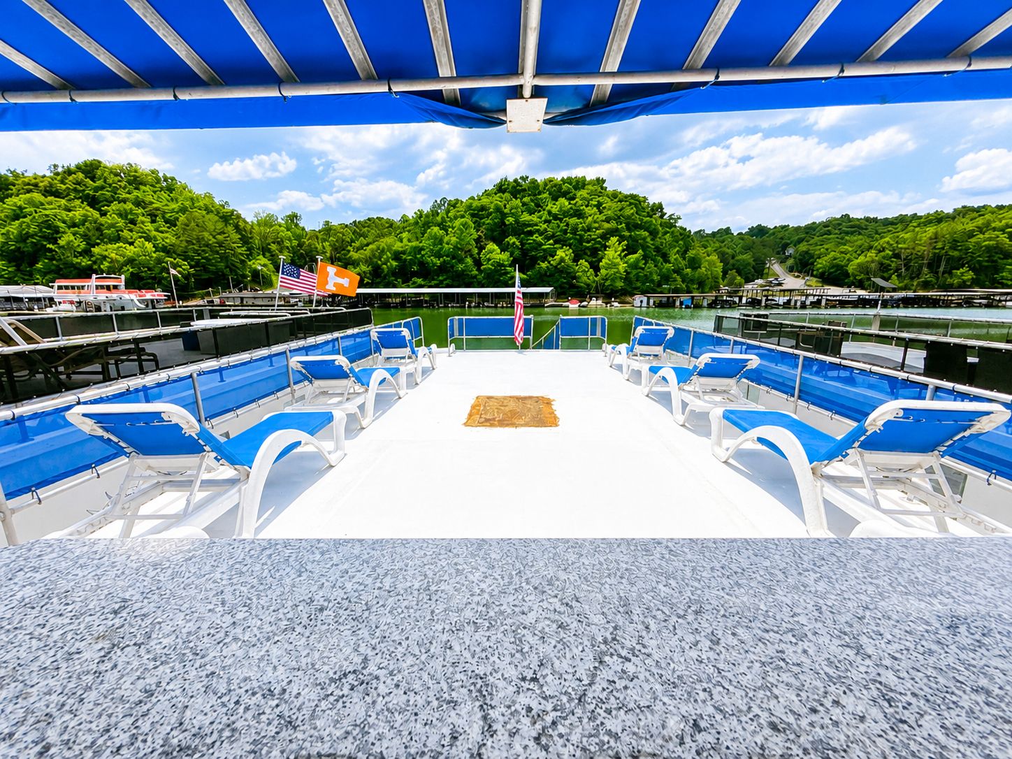 A houseboat with blue lounge chairs and awning, surrounded by a calm lake and green trees.