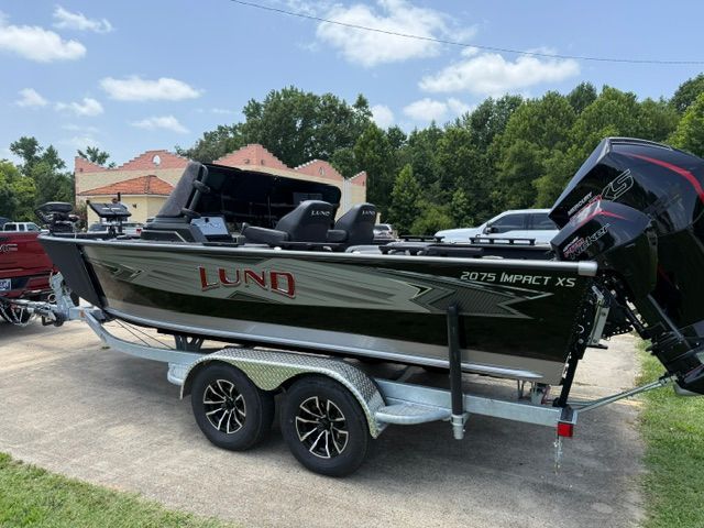 A black Lund 2075 Impact XS boat from 2025 sits on a trailer in a green setting.