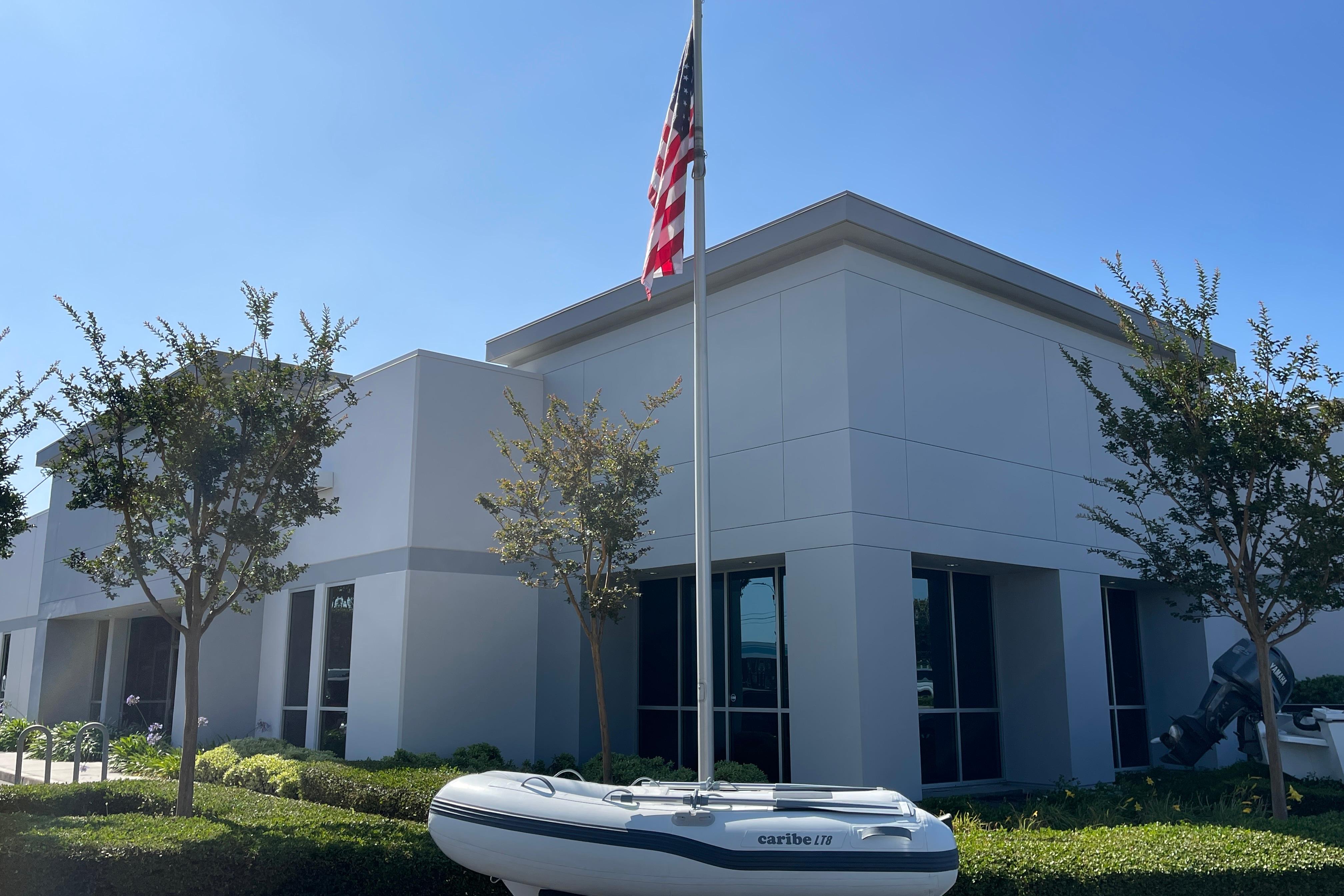 Modern building with flagpole, trees, and a 2025 Caribe L8 inflatable boat displayed in front.
